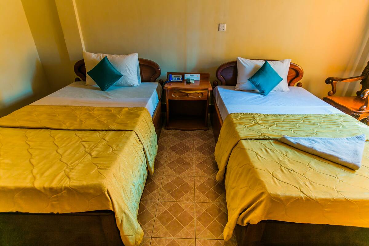 A hotel room with two single beds, each with a wooden headboard, a nightstand with a phone and a lamp, and a tiled floor.