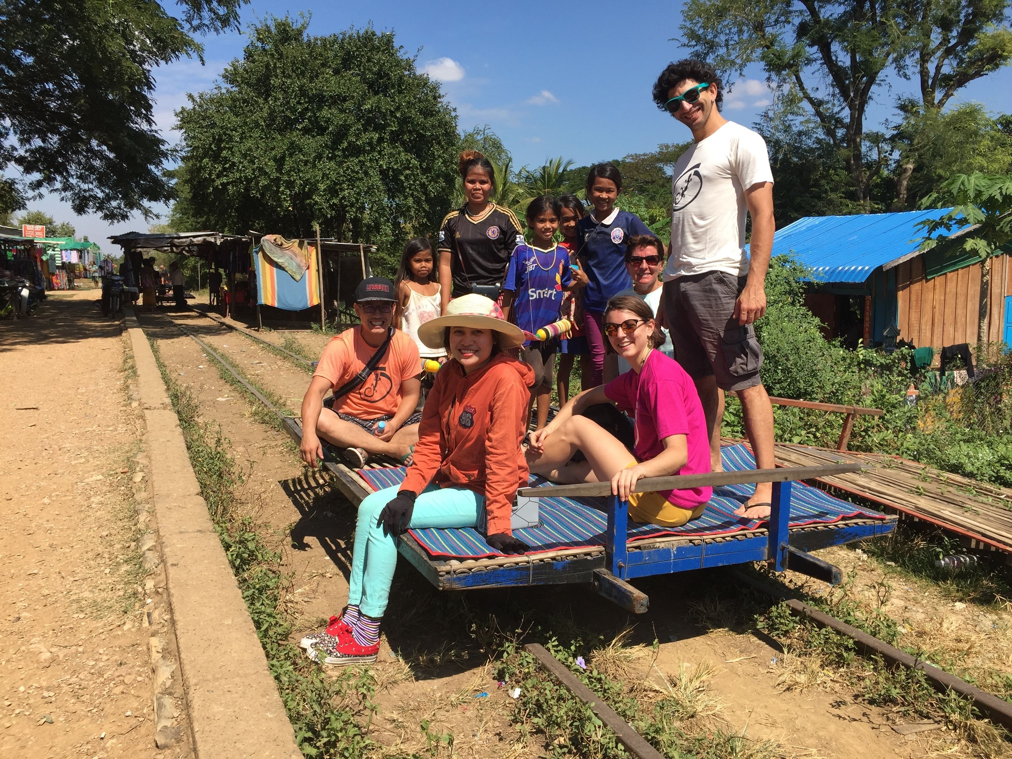 A group of people sitting on a wooden bench by a dirt path with a blue roof structure in the background.