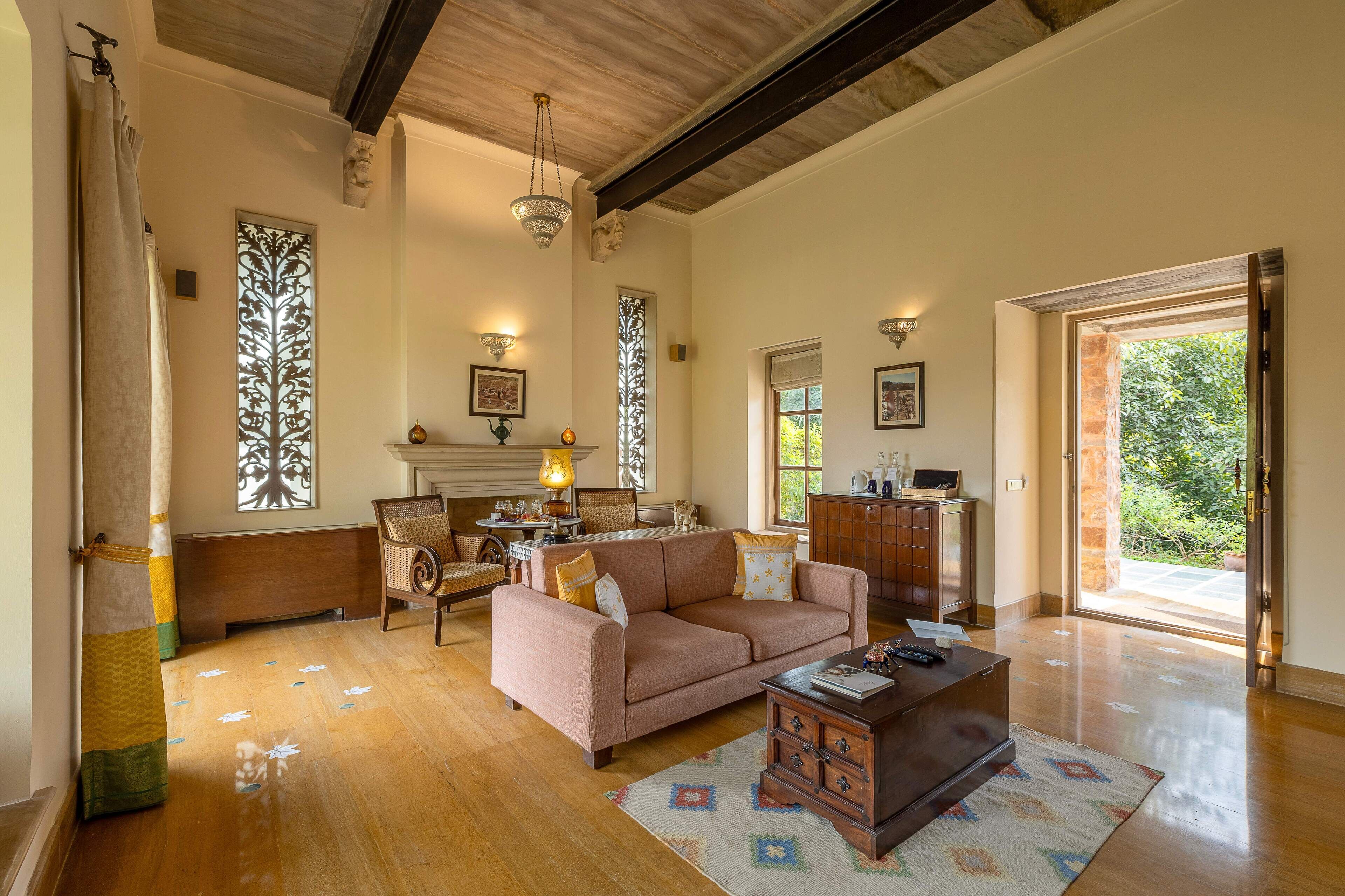 Amplia sala de estar con un sofá grande, una mesa de centro y una chimenea. Hay dos ventanas con contraventanas decorativas y vistas al exterior con vegetación.