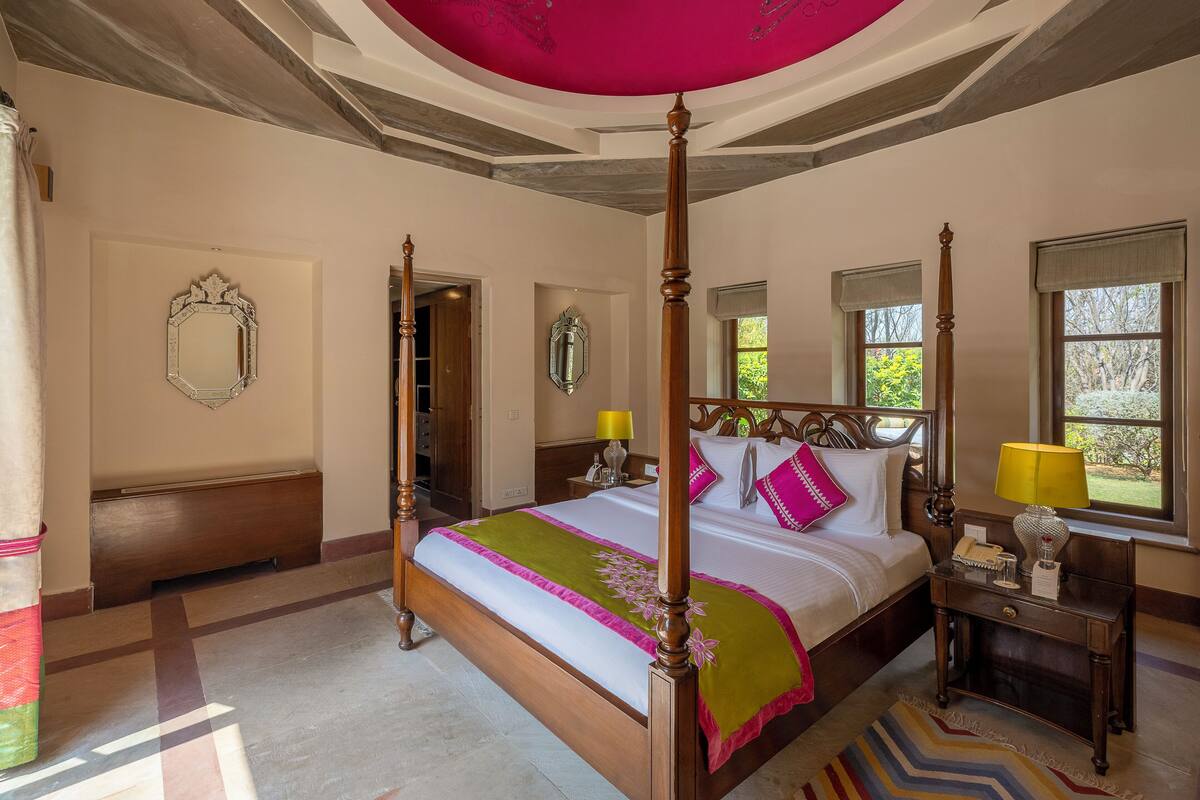 A four-poster bed with a green and pink bedspread, a wooden nightstand with a lamp, and a mirror on the wall.
