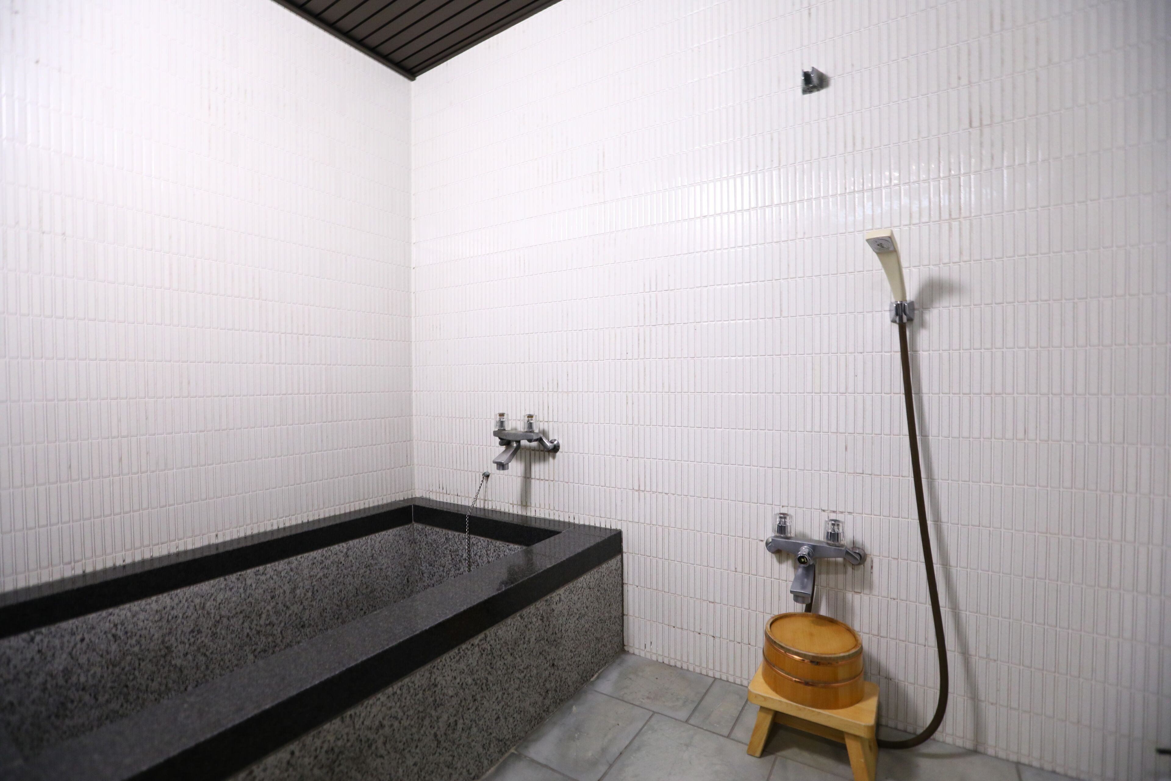 Un baño con una bañera rectangular, un taburete de madera y un cabezal de ducha.