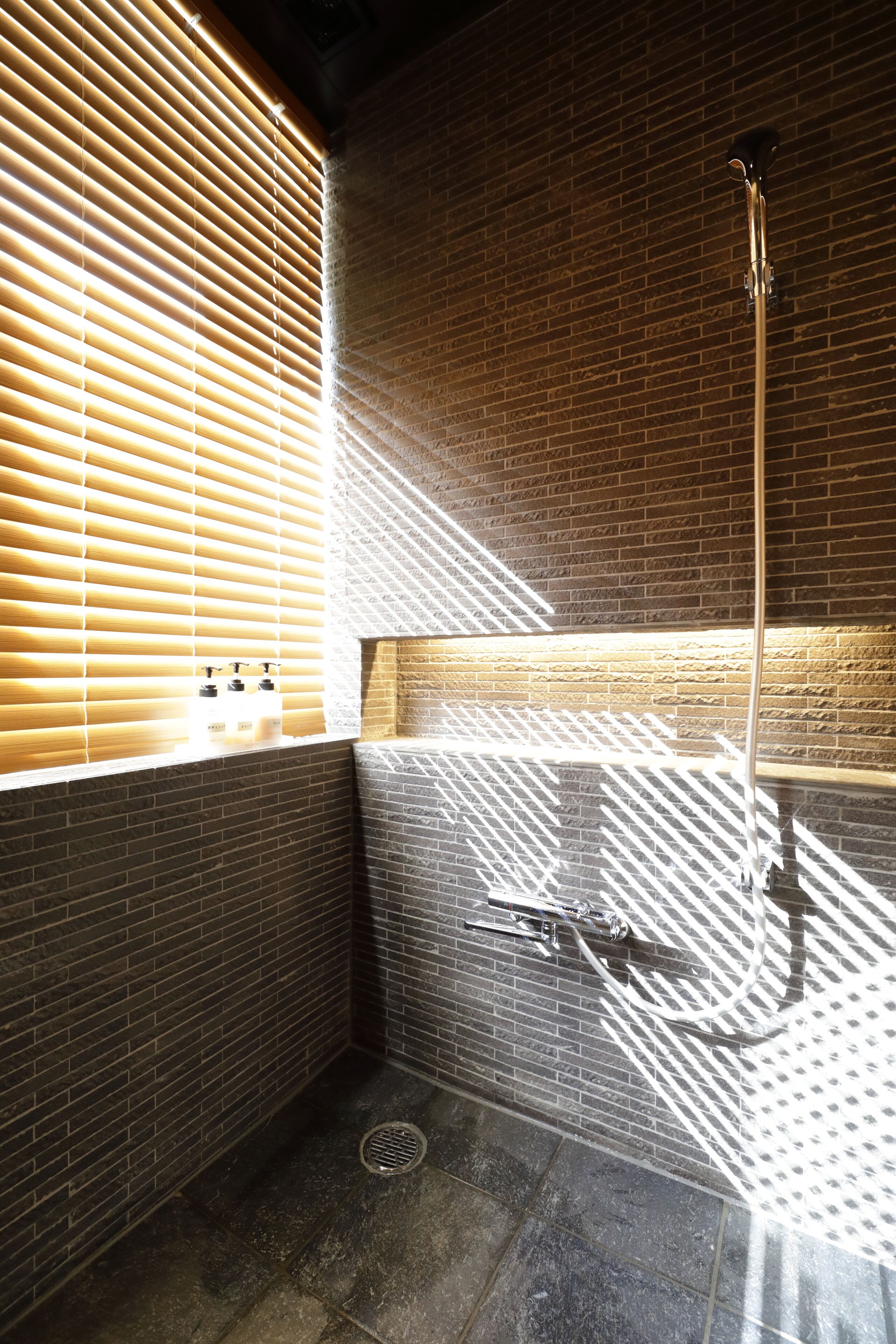 A bathroom with a shower, a window with blinds, and a brick wall.