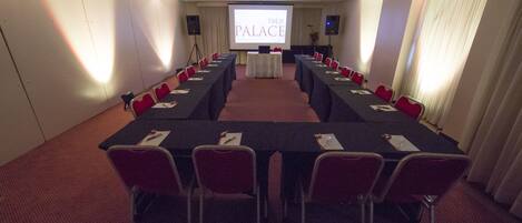 Un salón de conferencias con una larga mesa, sillas rojas y una pantalla de proyección que muestra "TSUE PALACE".