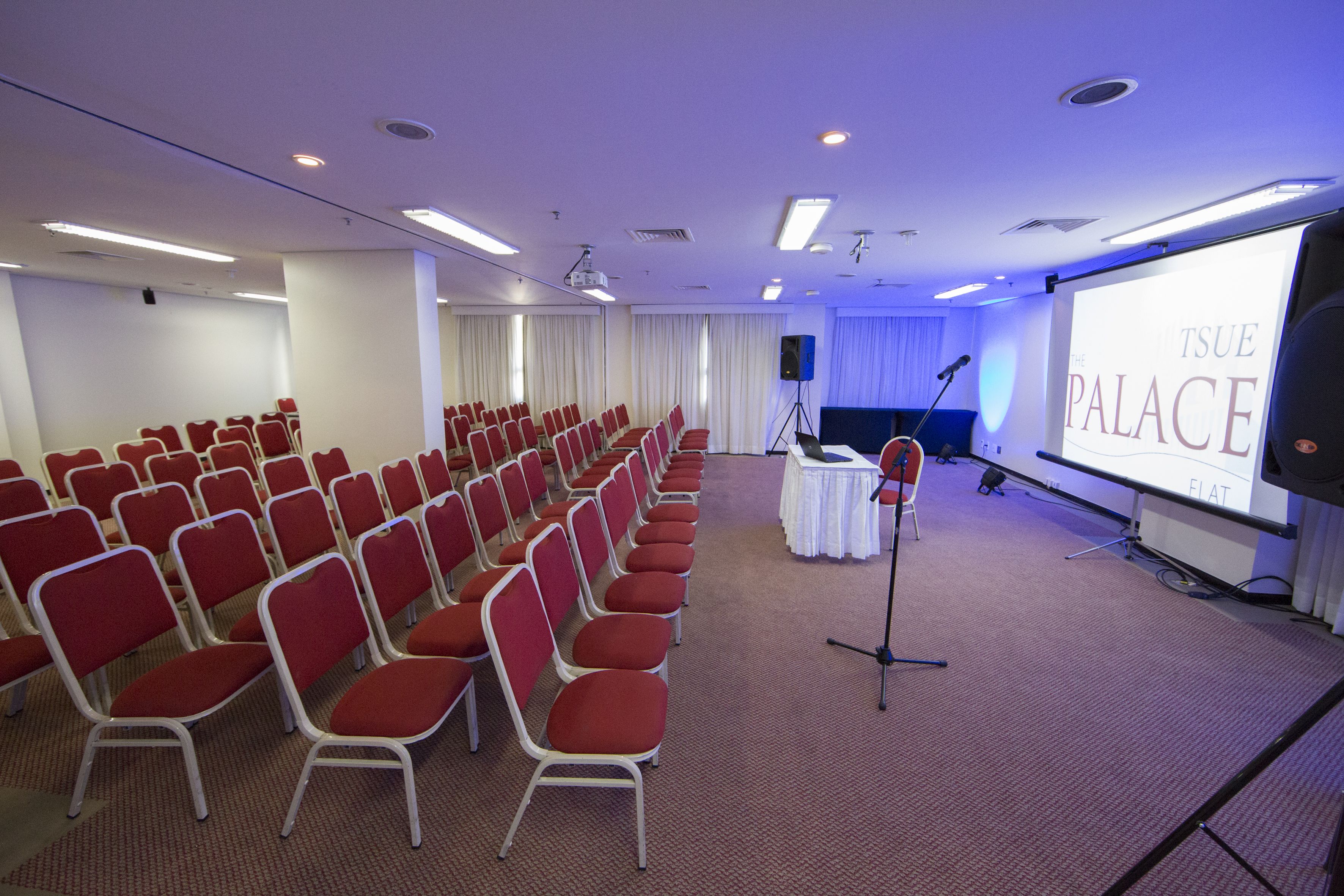 A conference room with rows of red chairs, a projector screen displaying "TSUE PALACE," and a stage with a microphone.