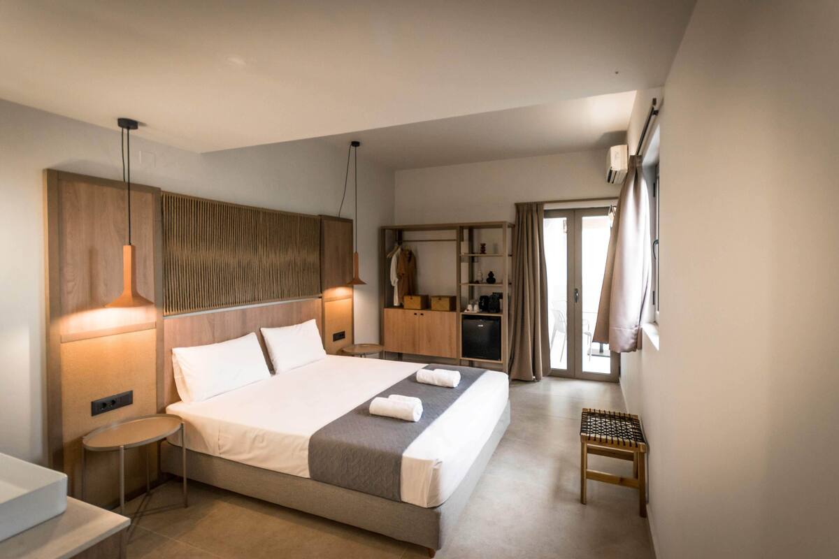 A modern hotel room with a large bed, bedside tables, a wardrobe, and a small seating area.
