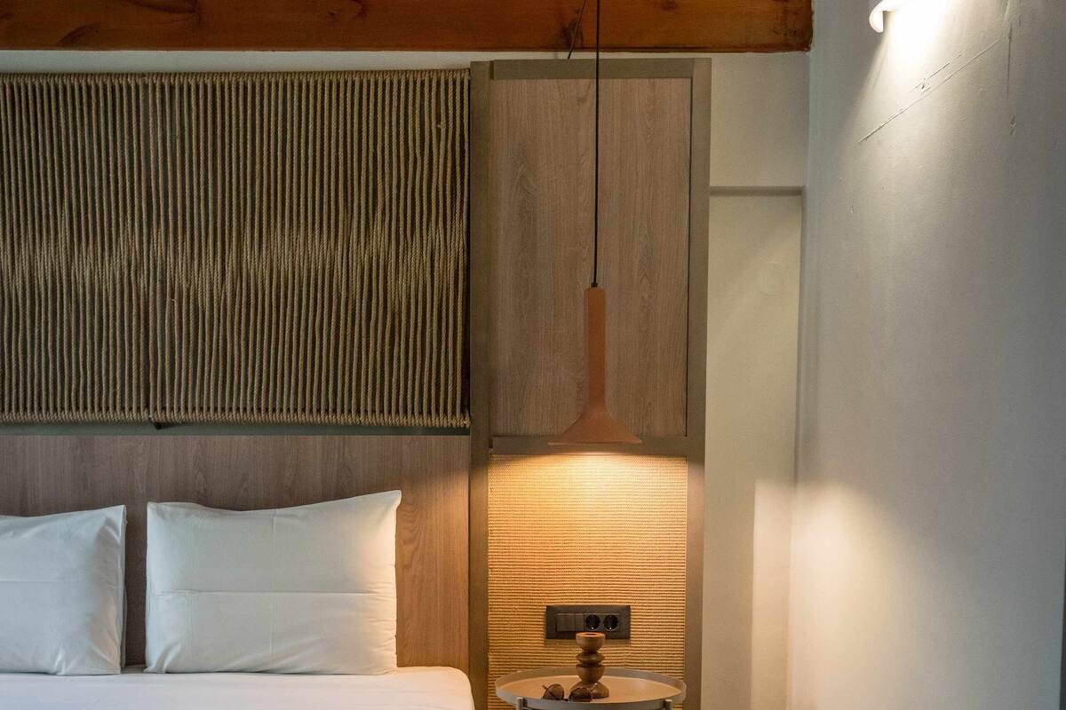 A hotel room with a wooden ceiling, a bed with white linens, a small table with a lamp, and a bench.