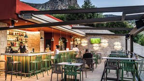 An outdoor dining area with green chairs and tables, a bar with a stone wall, and a mountain view. - Dream Village (Hersonissos)