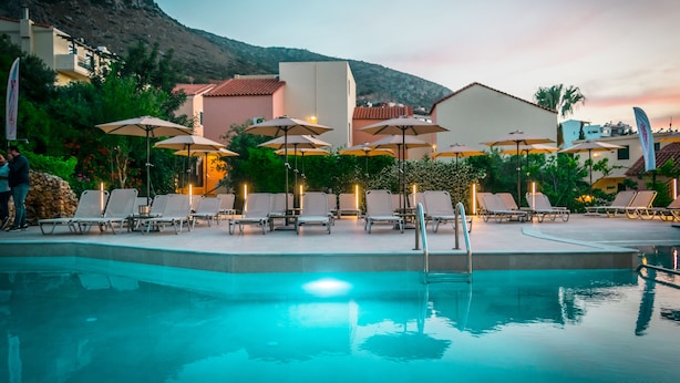 A pool area with lounge chairs and umbrellas, surrounded by buildings and a mountain backdrop. - Dream Village (Hersonissos)