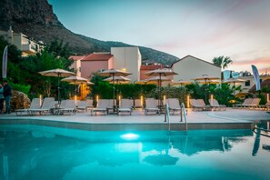 A pool area with lounge chairs and umbrellas, surrounded by buildings and a mountain backdrop. - Dream Village (Hersonissos)