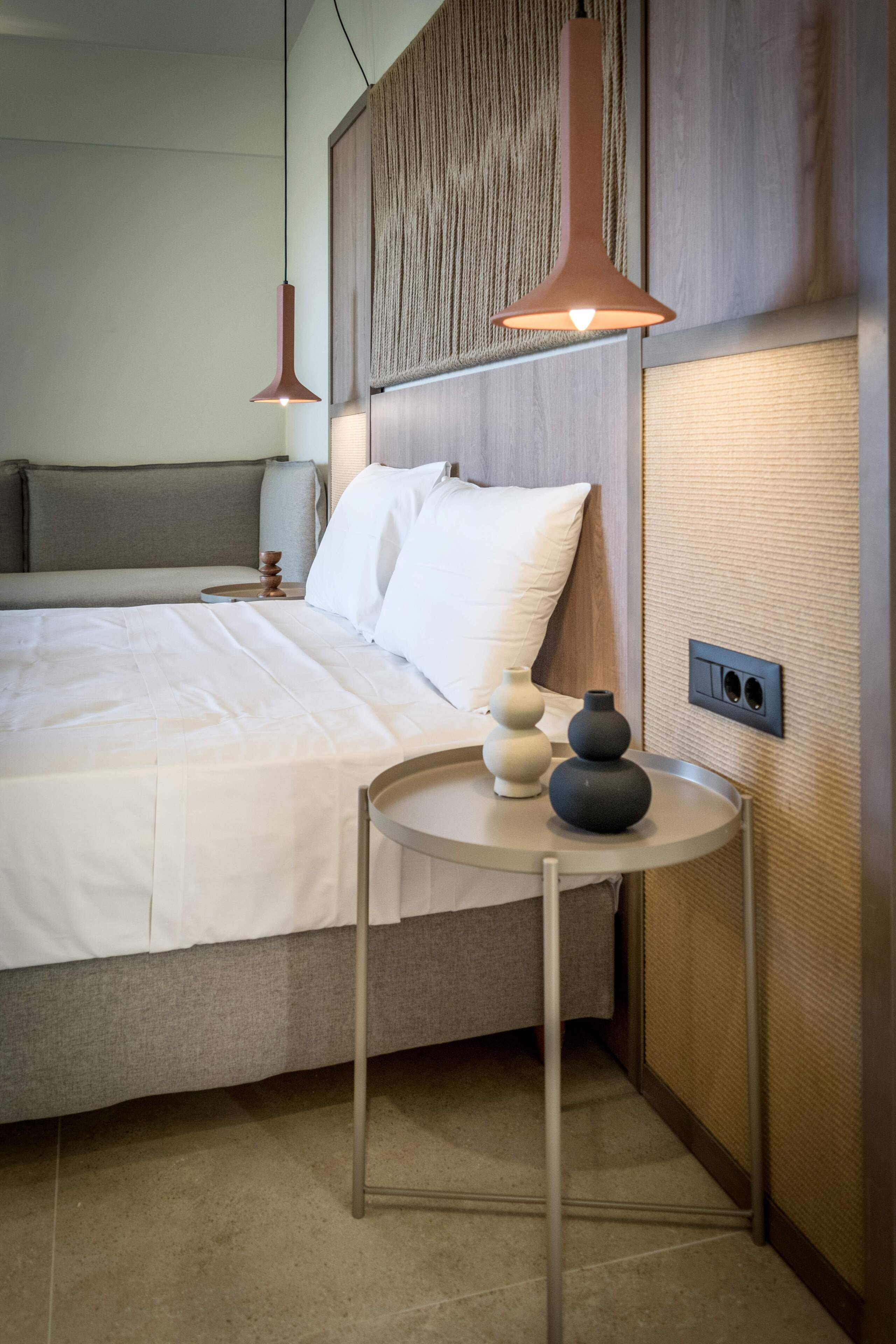 Un dormitorio moderno con una cama grande, dos lámparas colgantes, una mesita de noche con jarrones decorativos y un panel de pared con textura.