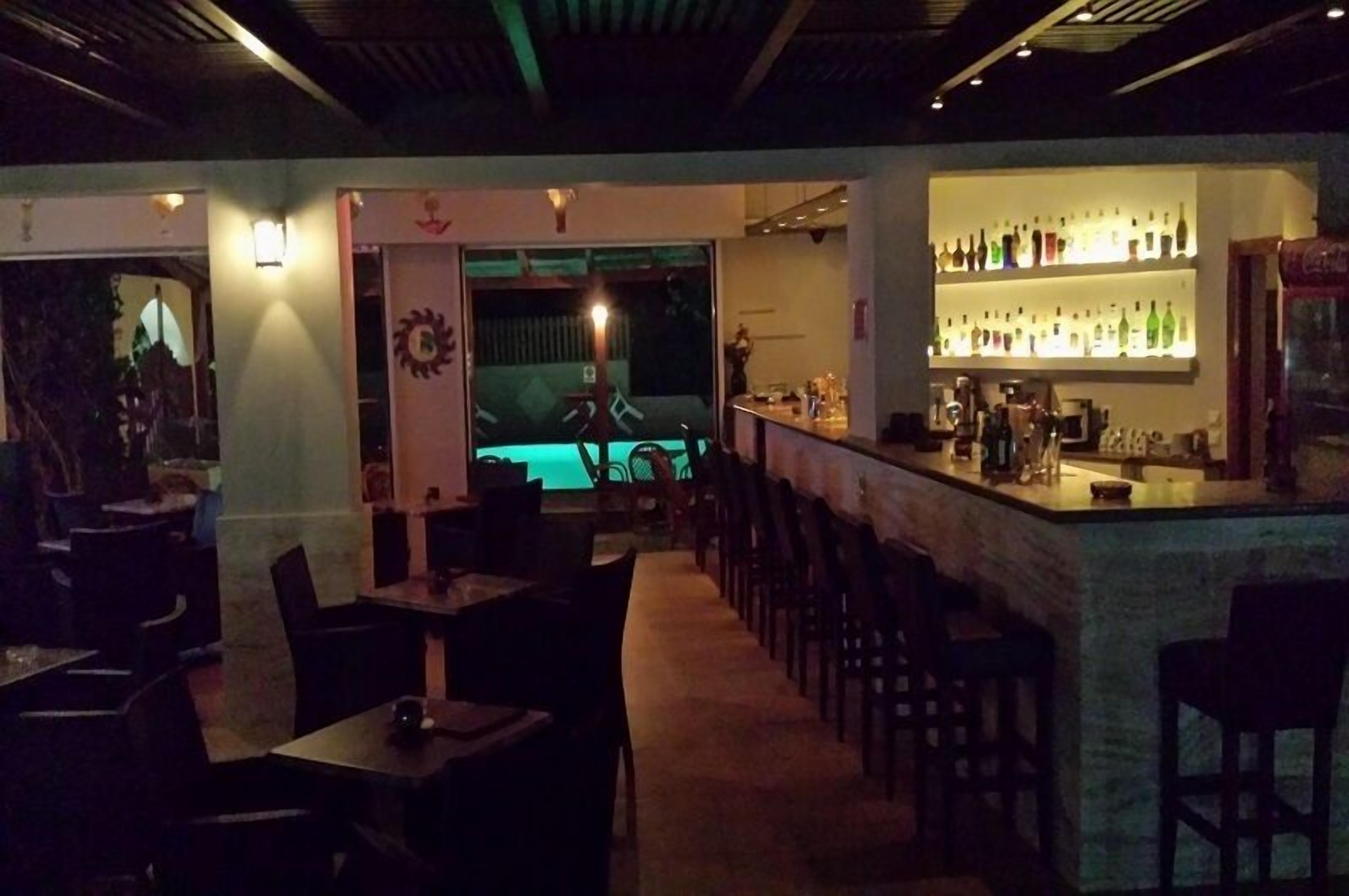 Zona de bar tenuemente iluminada con mesa de billar y barra mostrando varias botellas.