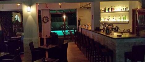 Zona de bar tenuemente iluminada con mesa de billar y barra mostrando varias botellas.