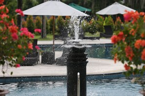 Ein Springbrunnen mit Wasserstrahlen in einem Poolbereich, umgeben von üppigem Grün und Außenmöbeln.