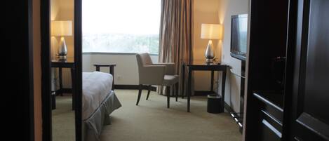 Quarto de hotel com cama, poltrona, escrivaninha, televisão e janela com cortinas.
