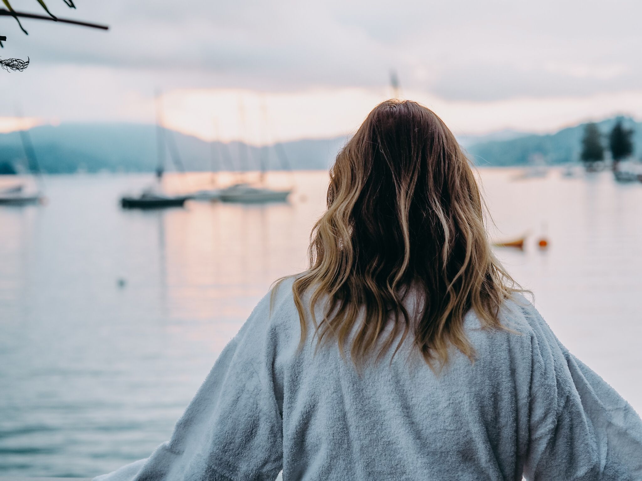 Une personne, vue de dos, avec de longs cheveux, est debout face à une étendue d’eau calme, où l’on aperçoit des bateaux et des montagnes au loin.