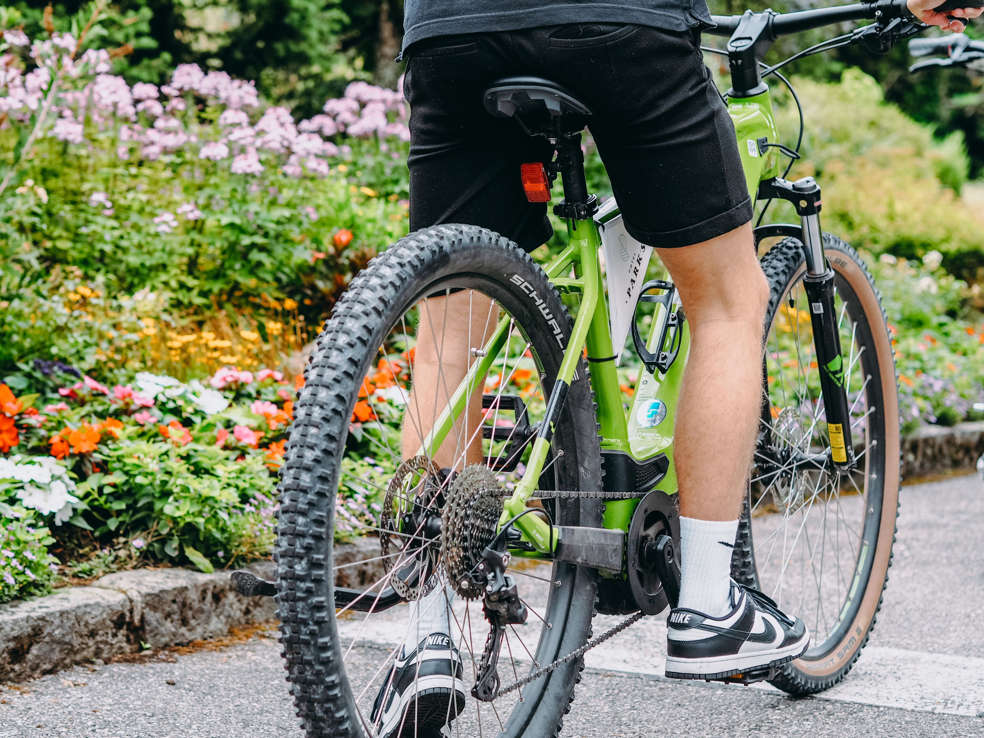 Een persoon staat naast een groene fiets met ketting en versnellingen, op een verharde ondergrond, met een tuin op de achtergrond.