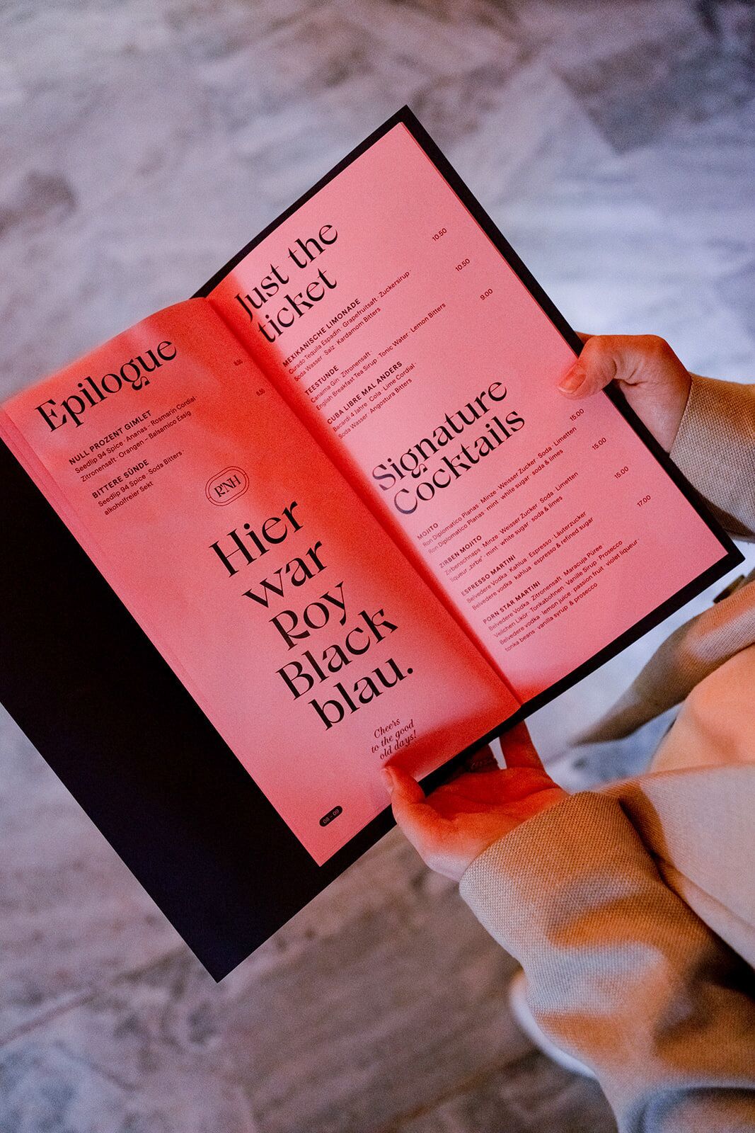 A person holding a menu with a red cover and black text, listing various drinks and prices.