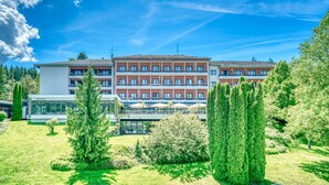 A multi-story hotel with a modern facade, a rooftop terrace with umbrellas, and a well-maintained garden with trees.