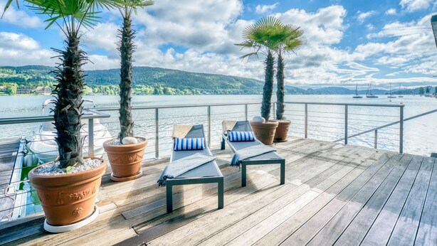A wooden deck with lounge chairs and potted palm trees overlooking a lake with boats.