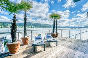 A wooden deck with lounge chairs and potted palm trees overlooking a lake with boats. - Hotel Parks (Velden am Wörther See)