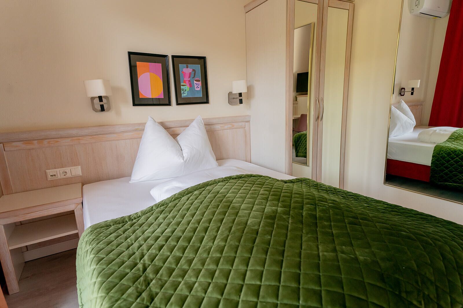 A hotel room with a bed, a bedside table, a mirror, and two wall-mounted picture frames.