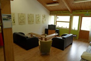 Een kamer met een houten plafond, een televisie op een standaard, een bank, een stoel en een tafel. Er hangen ingelijste botanische prints aan de muur en een raam met uitzicht op groen buiten.