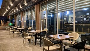 Un área de café al aire libre con mesas, sillas, ventanas amplias y una barra visible en el interior.