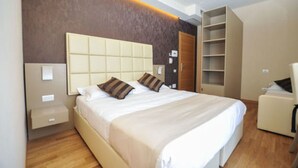 Moderni hotellihuone, jossa on suuri sänky, yöpöytä, vaatekaappi ja oleskelualue.
