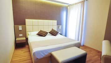 Quarto de hotel com uma cama grande, mesa de cabeceira, uma cadeira e uma janela com cortinas.