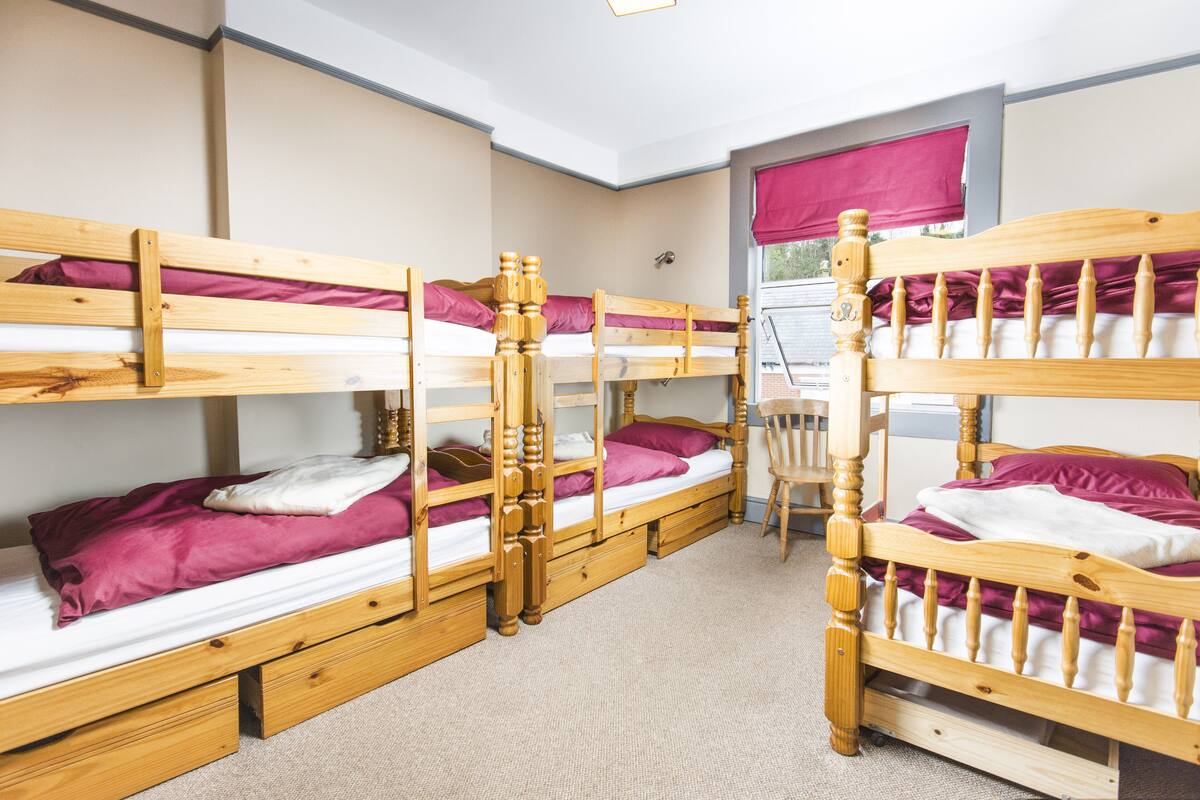 A room with four bunk beds, each with wooden frames and purple bedding. There is a window with a red blind, and a chair is visible in the corner.