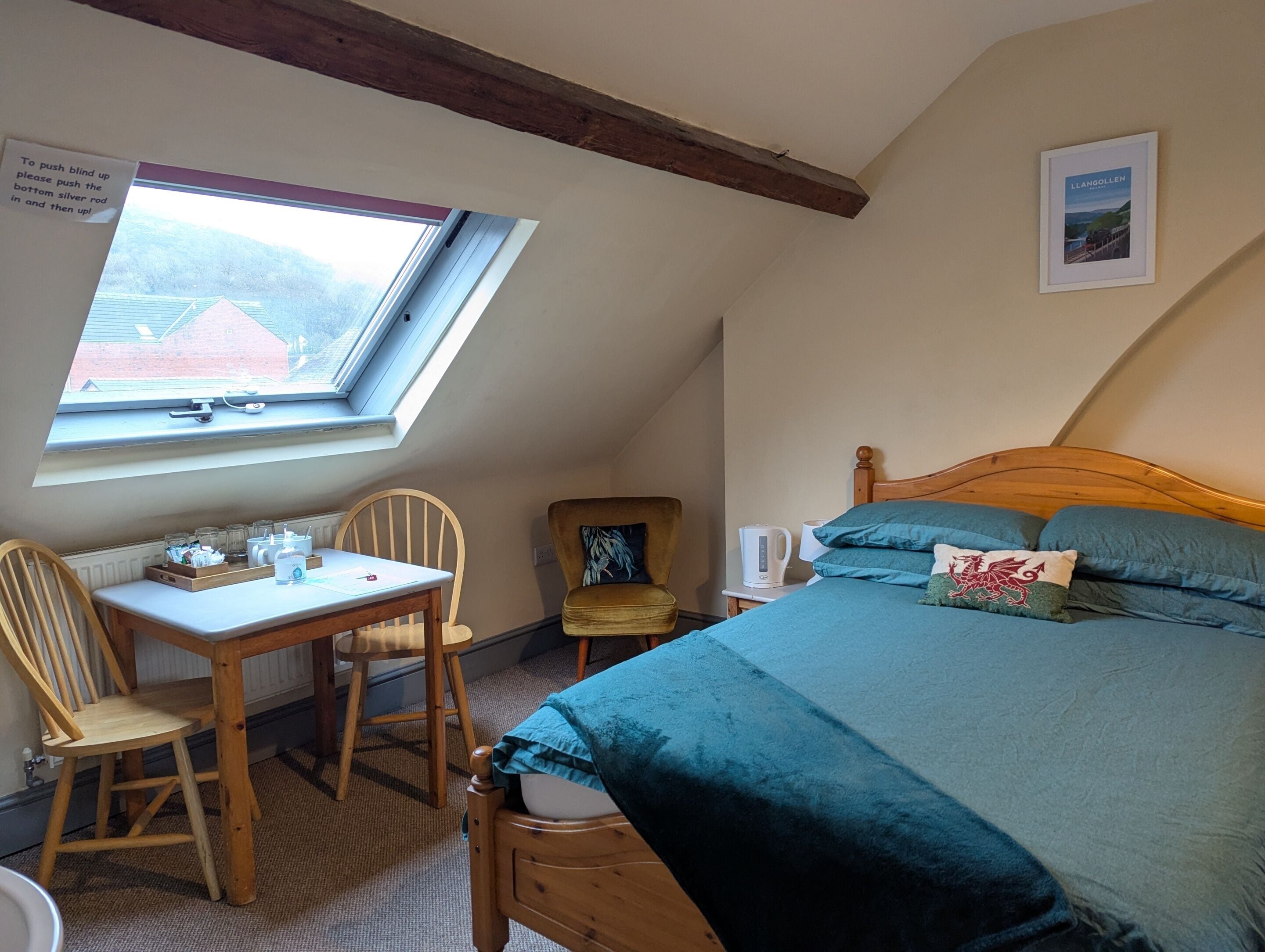 Una camera accogliente con soffitto spiovente, un letto di legno, una scrivania con sedie e una finestra con vista su una casa.