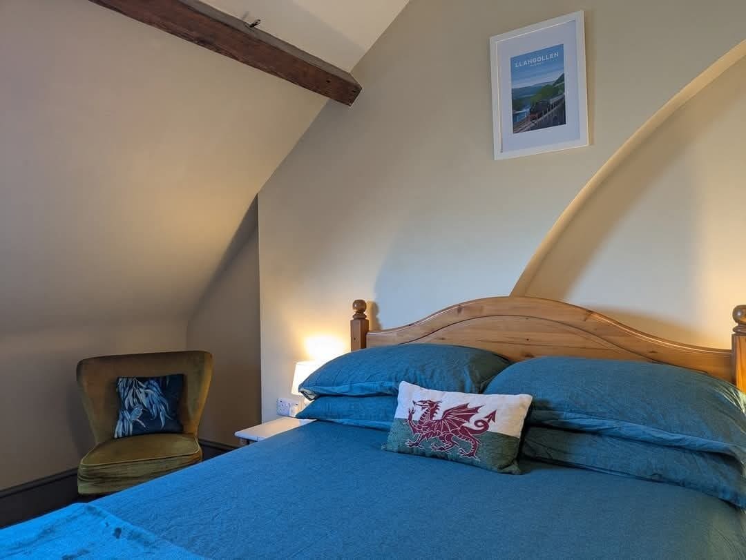 Un dormitorio con cabecera de madera, cama con ropa de cama azul, una silla con cojín estampado y un cuadro en la pared.