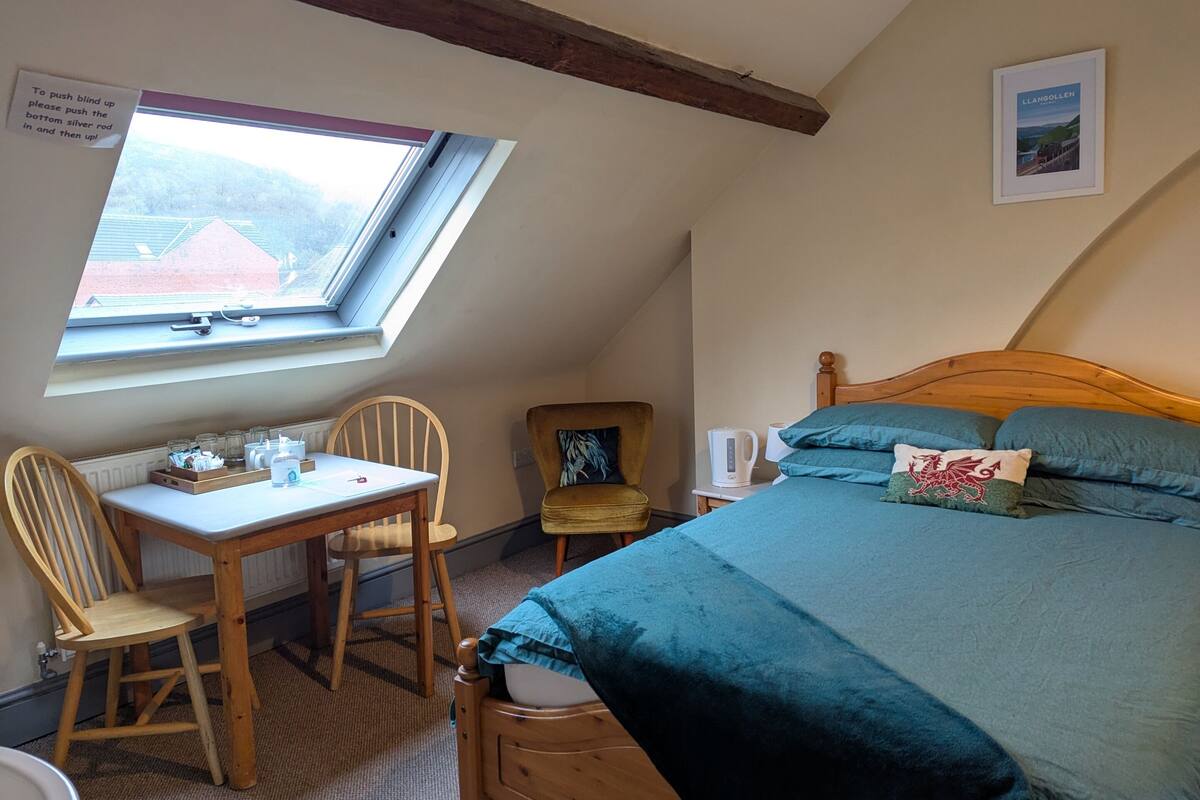 A cozy bedroom with a wooden bed, a desk with a chair, a window, and a picture on the wall.