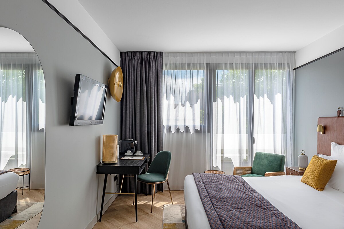 Una habitación de hotel con cama, escritorio con silla, televisión y ventana con cortinas.