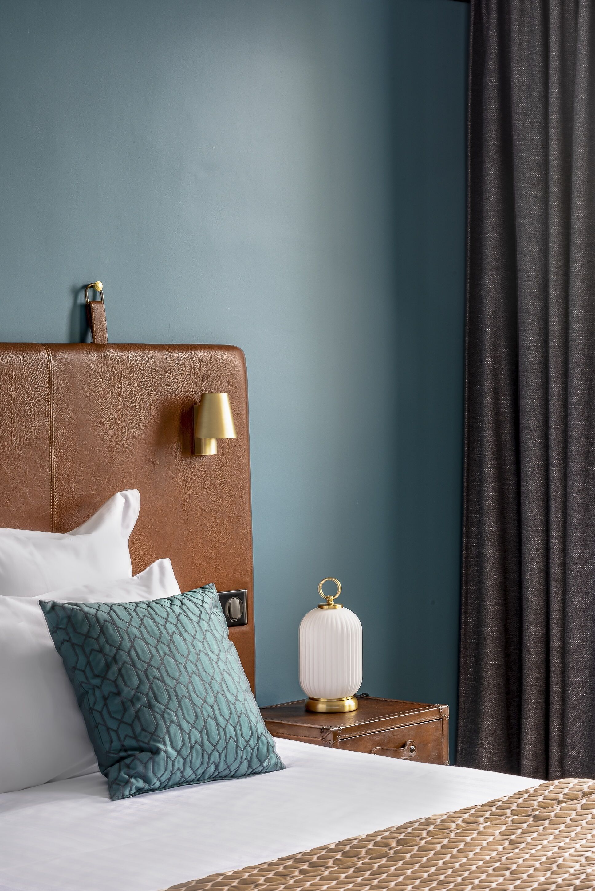 Una cama bien hecha con un cabecero marrón, una almohada blanca, una almohada estampada color turquesa y una lámpara blanca sobre una mesita de noche de madera.