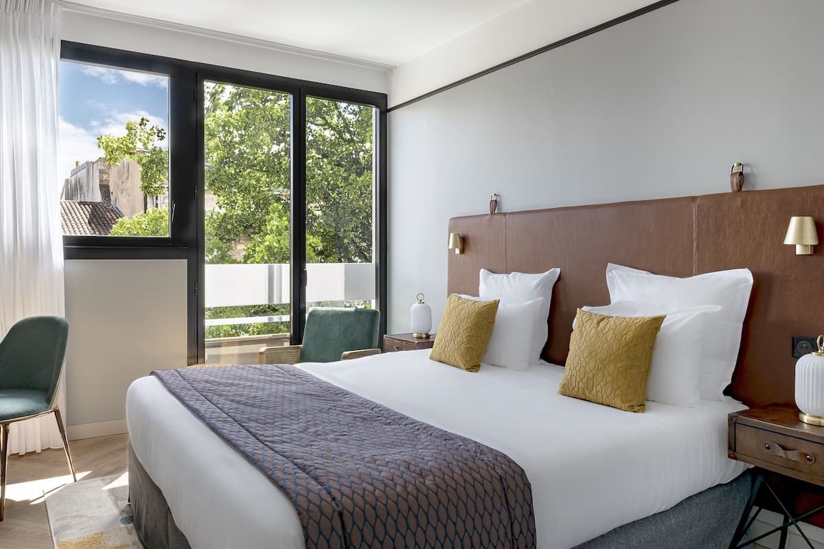 A modern hotel room with a large bed, a green chair, a nightstand, and a view of the outdoors.