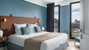 Habitación de hotel moderna con una cama grande, una mesita de noche, una lámpara y vistas de la ciudad a través de un ventanal amplio.