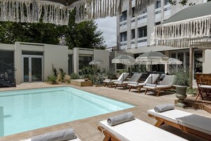 Zona de piscina con sillas de descanso y sombrillas, rodeada por un edificio con balcones y un jardín.