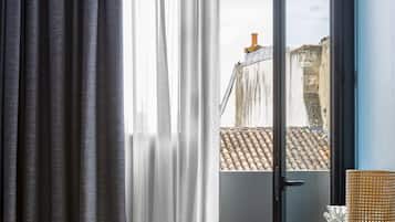 A room with a view of buildings through a window, a chair, a table with a vase, and a hanging light fixture.