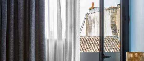 A room with a view of buildings through a window, a chair, a table with a vase, and a hanging light fixture.