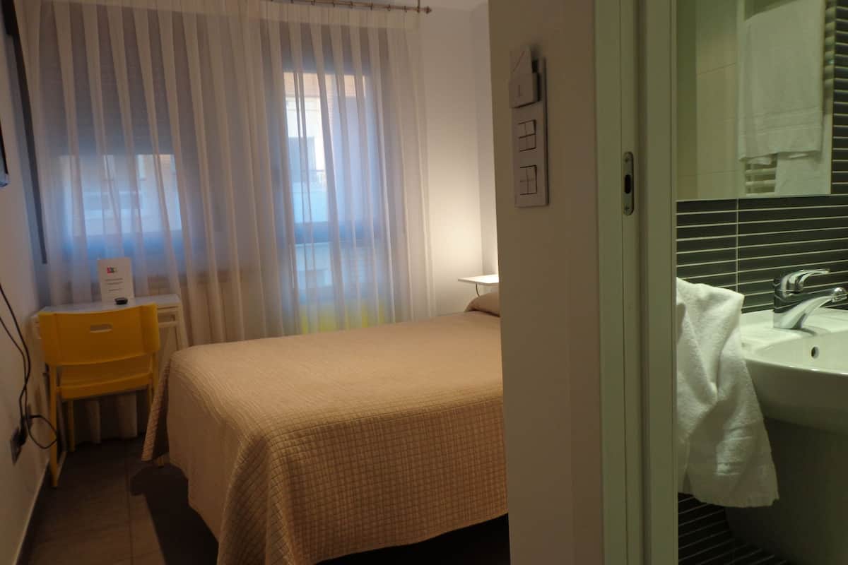 Habitación de hotel con cama, una silla, escritorio, ventana con cortinas translúcidas y baño con lavabo y espejo.