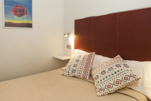 Una cama bien hecha con almohadillas estampadas, un cabecero de madera, una mesita de noche con lámpara y un cuadro en la pared.