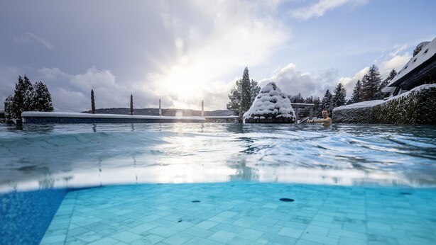 A clear blue pool with a tiled floor, surrounded by snow-covered trees and a building with a sloped roof.
