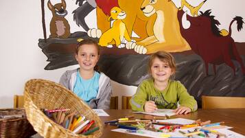 Deux enfants sont assis à une table, devant du matériel de dessin, et un grand décor mural représentant des personnages de dessins animés se trouve à l’arrière-plan.