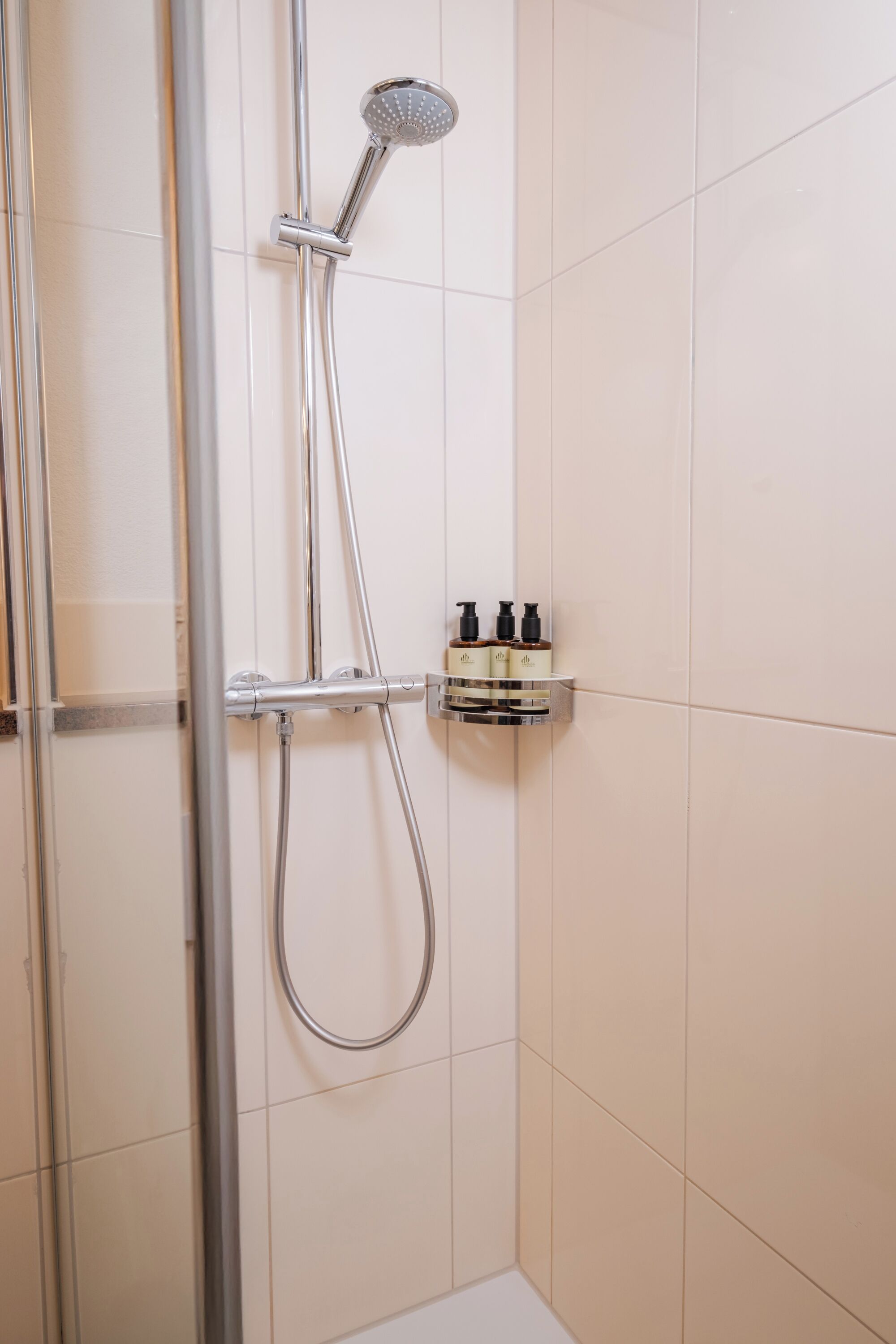 A shower with a handheld showerhead and a wall-mounted faucet, a small shelf with toiletries, and a tiled wall.