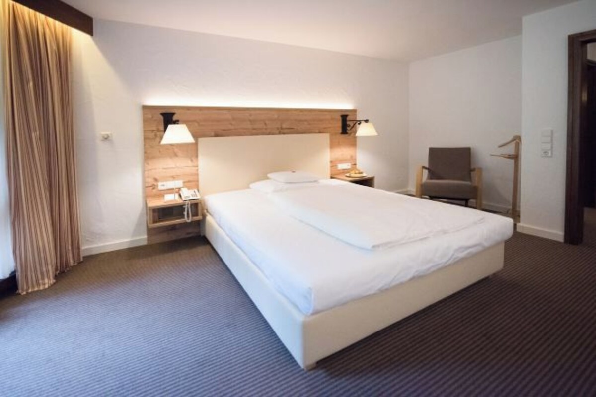 Una habitación de hotel con una cama grande, dos mesitas de noche con lámparas, una silla y un cabecero de madera.