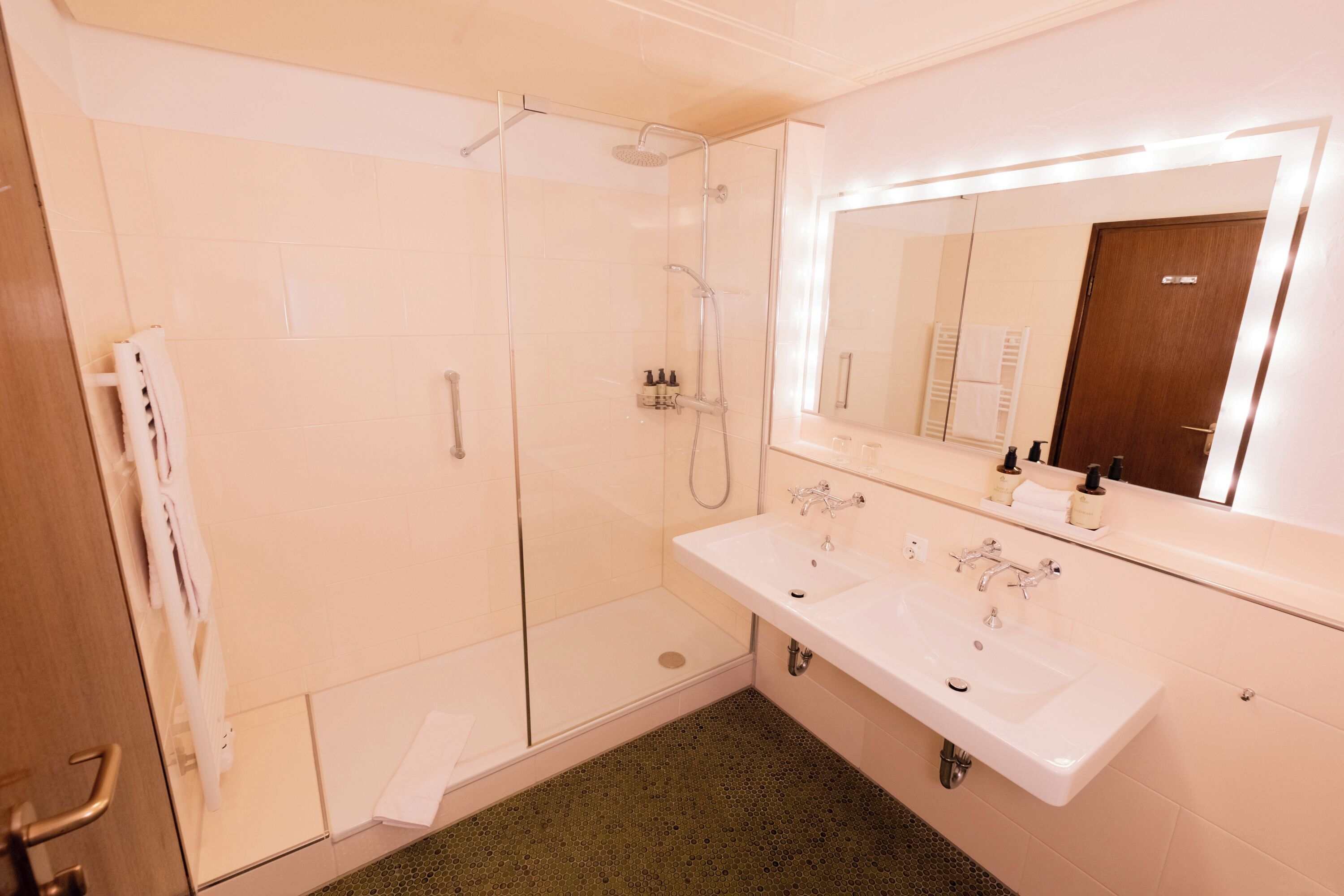 Un baño moderno con ducha de vidrio, lavamanos doble y un espejo grande con luces.