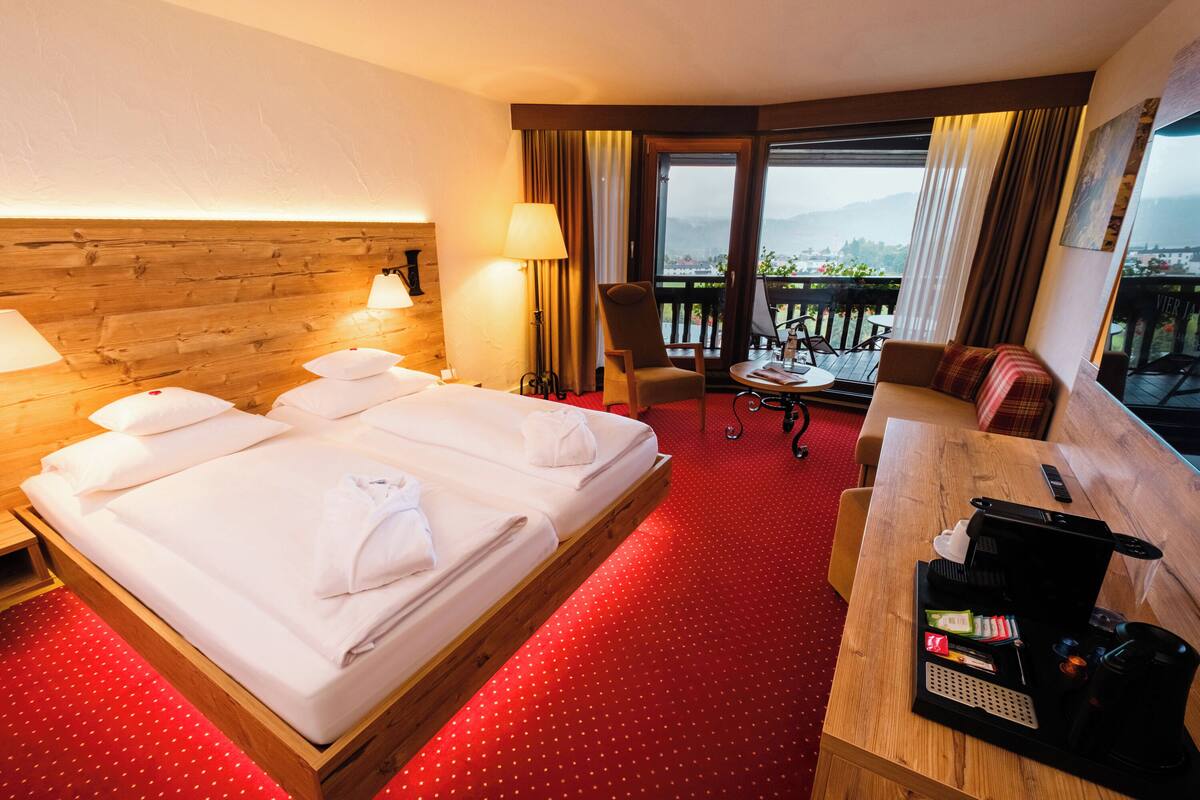 A hotel room with a bed, a desk with a coffee maker, a chair, and a view of the outdoors.