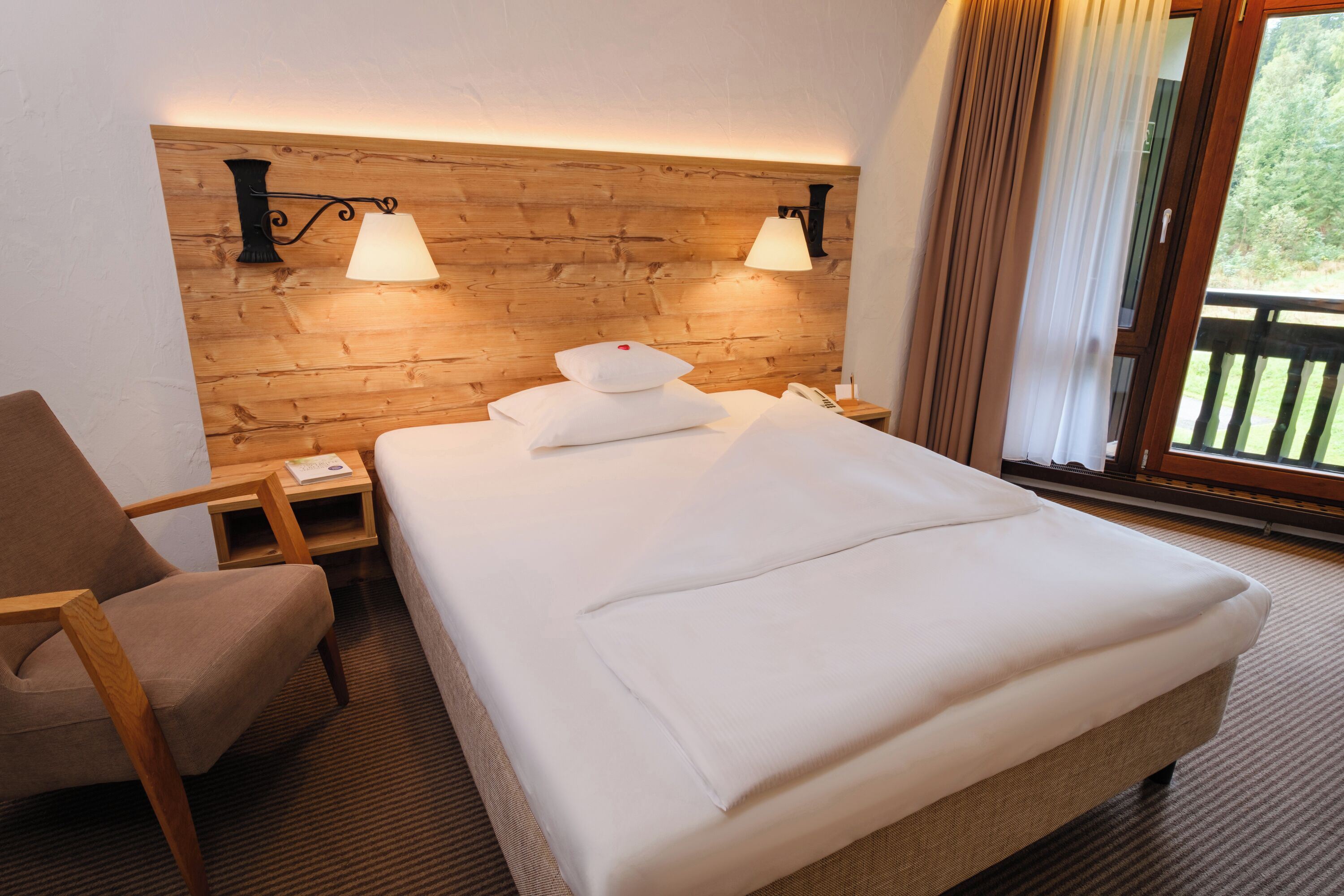 Hotelkamer met houten hoofdeinde, twee bedlampen, een bed met witte lakens, een houten stoel, een nachtkastje en uitzicht door een raam met gordijnen.