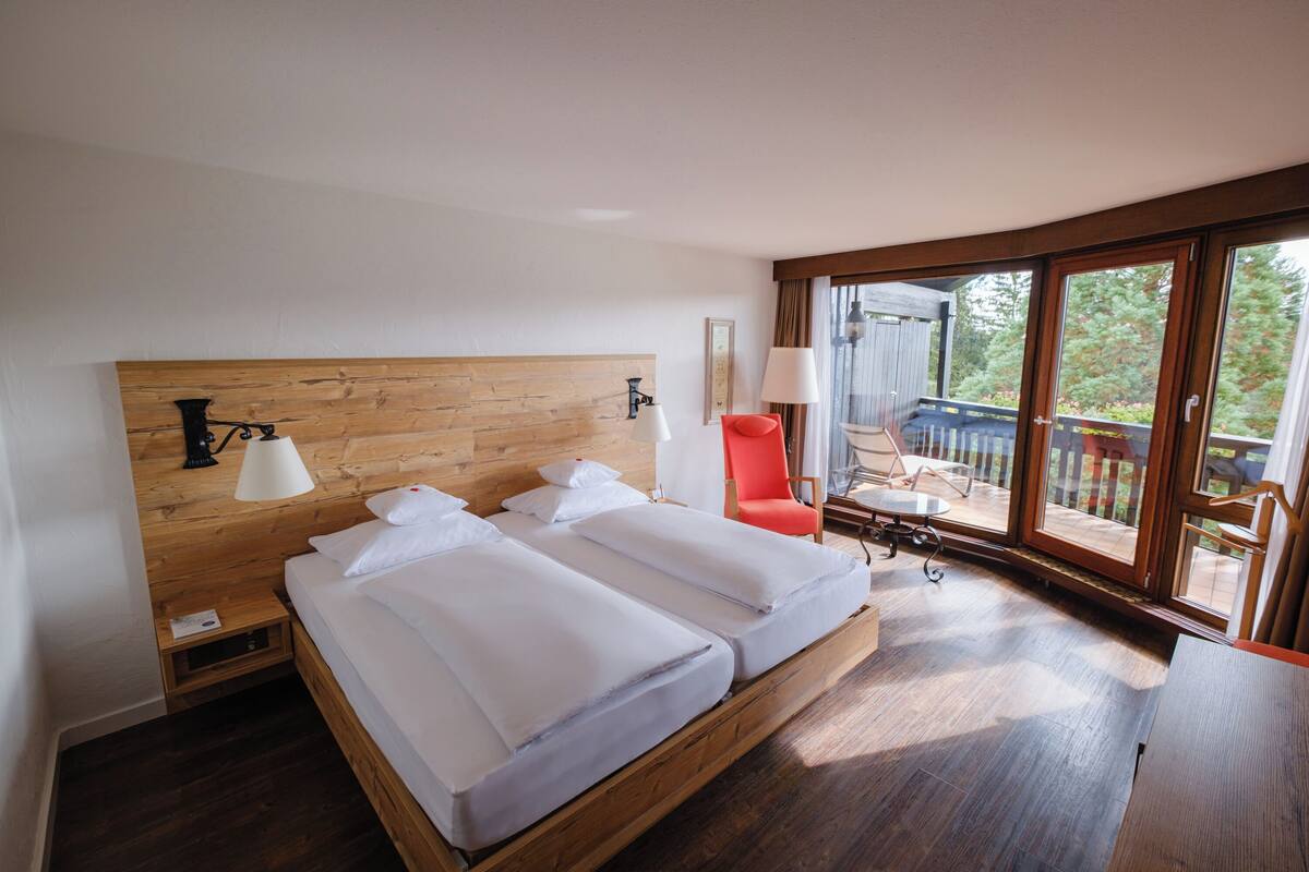 A bedroom with a wooden headboard, two beds, a nightstand, a red chair, and a balcony with a view.